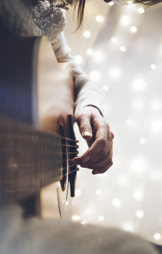 Hipster girl playing guitar in a home atmosphere, person studying on musical instrument on glow bokeh Christmas illumination, female hands using guitar in holiday on relax glitter decoration, blur