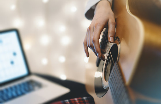 Hipster girl playing guitar in home atmosphere, person studying on musical instrument and notes in laptop on background glow bokeh Christmas illumination, holiday on relax glitter decoration, blur