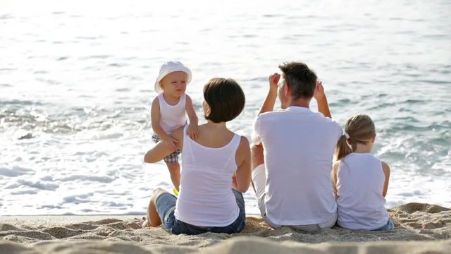 man and woman with two kids sitting with back to camera on sandy beach
