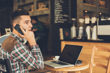 Young man talking on the phone with a laptop on table in cafe