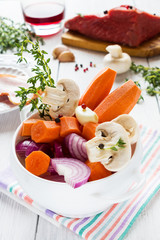 Ingredients for Boeuf Bourguignon, carrot, onion, mushrooms, bacon, wine, beef, garlic, thyme. White bowl, wooden table.