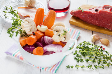 Ingredients for Boeuf Bourguignon, carrot, onion, mushrooms, bacon, wine, beef, garlic, thyme. White bowl, wooden table.