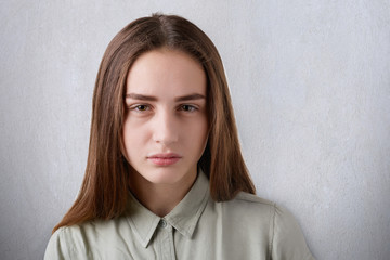 Obraz premium A close-up of a beautiful young girl with shining eyes and straight long dark hair having a sombre face looking straight in the camera. A portrait of cute girl on white background falling to thinking
