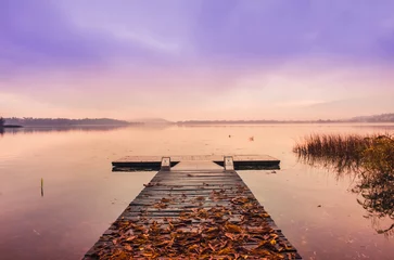 Wall murals Pier Wooden pier with leaves on the quiet lake at sunrise in autumn