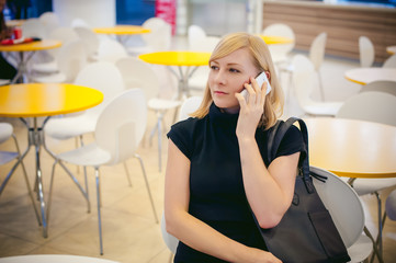 blonde girl in a black dress talking on the phone while sitting at a table in a cafe