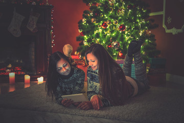 Obraz na płótnie Canvas Young female friends lying on rug beside Christmas tree and looking at the tablet. Beautiful new year room interior