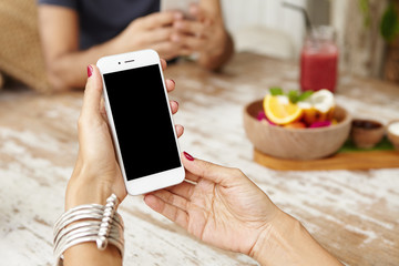 White smart phone with blank copy space screen in woman's hands over cafe table. Cropped shot of female using wi-fi, checking e-mail on her electronic gadget, sitting at cafeteria with her husband