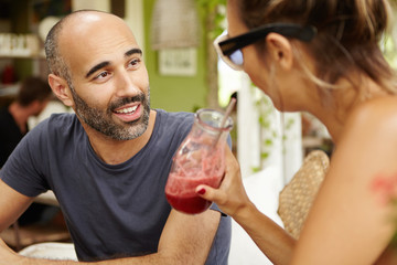 Handsome middle-aged man with stubble sitting at sidewalk restaurant, having nice conversation with attractive female who is drinking smoothie. Two tourists talking over lunch at cafe during holidays