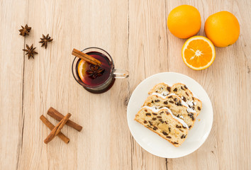A of glass of Mulled Wine and slices of Christstollen surrounded by fresh oranges, cinnamon sticks and star anise. Shot from directly above on light brown wooden background.