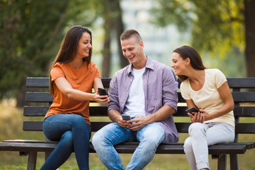 Three friends are sitting on bench in park and using smartphones. 