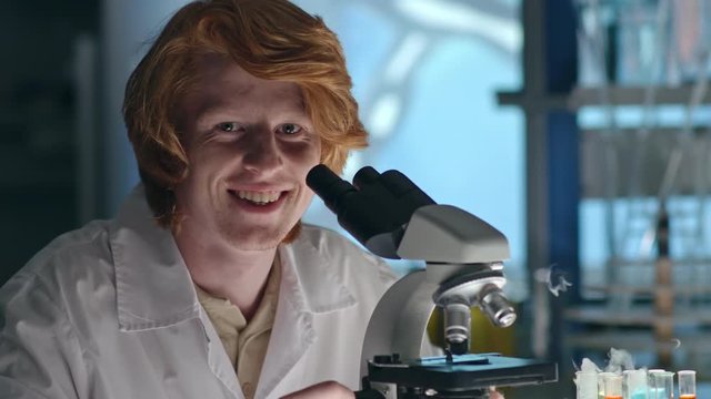 Closeup of young redhead chemistry student looking through microscope and then looking at the camera and smiling