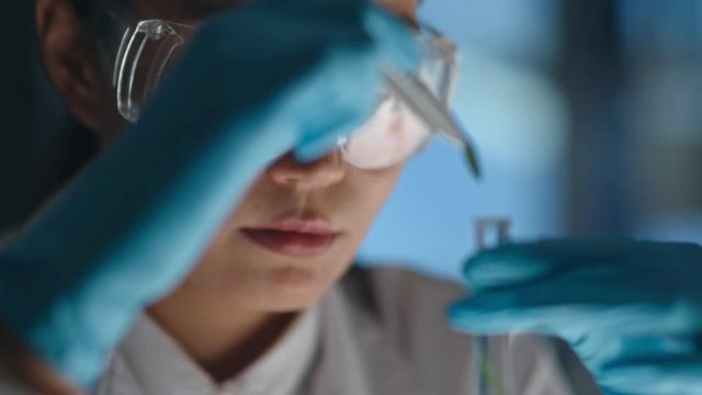 Closeup of woman in protective glassware putting green leaves into glass tube with tweezers and looking at reaction