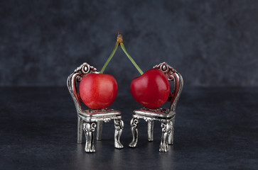 Couple of red cherries placed on beautiful silver vintage chairs on dark background with copy space. Relationship concept.