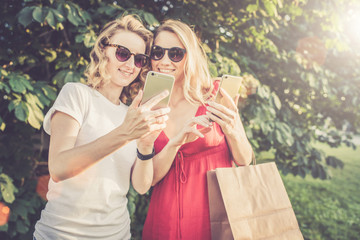 Obraz premium Summer sunny day. Two young women with blond hair laughing looking at smartphone screen. The girl in a red dress holding a shopping bag. Girls use digital gadgets. Selective focus, film effect.