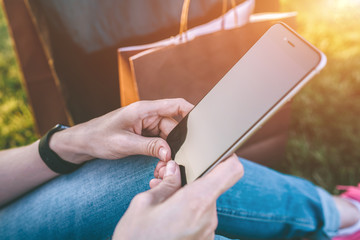 Summer evening.Close-up of smartphone with blank screen in hands of young woman,dressed in blue jeans,sitting outside on lawn.Girl resting after shopping and using smartphone.Nearby are shopping bags.