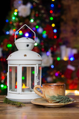 Christmas Decorations with small cup of coffee, lamp with candle, fir branch on wooden table against lights background