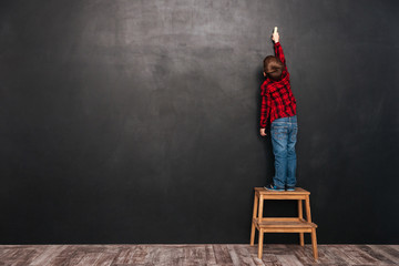 Child standing on stool near blackboard and drawing at board