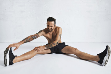 Obraz premium Bearded attractive tattooed male athlete stretches his legs, sitting in white room and smling on camera