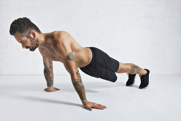 Obraz premium Strong brutal tattoed and ripped male athlete shows how to do pseudo push-ups, isolated on white spacious room, top position