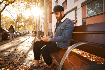 Bearded hip young man on a sunlit autumn city street sitting on a bench looking at his smartphone screen