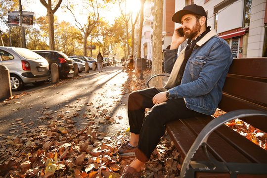 Attractive young man with a beard dressed in denim jacket, black trucker hat and black trousers sitting on a wooden bench in an autumn city street talking on the phone