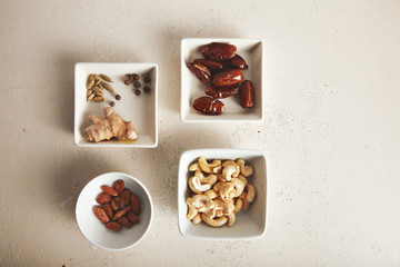 Various nuts, dried fruits and spices beautifully arranged in small white bowls of different shapes on rough white surface, top view