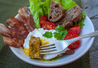 Classic hearty breakfast. Fried egg, bacon, vegetables (lettuce, tomato), greens and bread made from whole wheat flour.