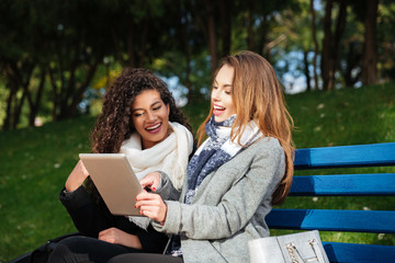 Young beautiful ladies sitting on bench and looking at tablet