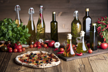 Delicious, tasty and fresh, rustic Italian pizza, served on wooden table. Bunch of tomatoes, chili, basil, garlic in background. Bowls with different kinds of pepper. Many bottles of olive oil