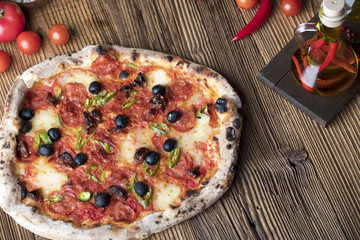 Delicious, tasty and fresh, rustic Italian pizza, served on wooden table. Bunch of tomatoes, chili, basil, garlic in background. Bowls with different kinds of pepper. Many bottles of olive oil