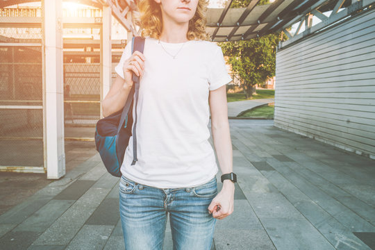 Cropped image. Front view. Young woman with a backpack, wearing a white T-shirt and blue jeans, standing outdoors. Summer sunny evening.