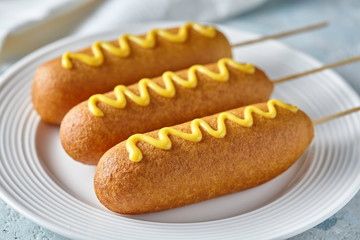 Corn dog traditional American corndog street junk food fried hotdog meat wiener with mustard on top snack treat in a layer of cornmeal batter on stick unhealthy eating on rustic table.