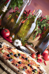 Delicious, tasty and fresh, rustic Italian pizza, served on wooden table. Bunch of tomatoes, chili, basil, garlic in background. Bowls with different kinds of pepper. Many bottles of olive oil