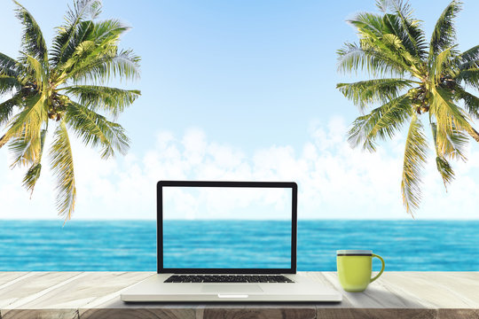 A laptop and a coffee cup on wood table in the sea beach with coconut tree.
