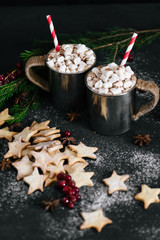 Hot cacao with marshmallows in metal cups, star shaped cookies and red berries on black table