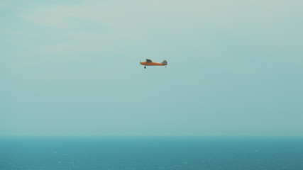 Fototapeta premium Orange one engine prop plane with teal stripes flying above misty ocean in Rio de Janeiro, Brazil, bright sunny day with clear sky