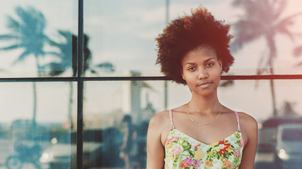 Portrait of young beautiful cute mixed brazilian teen girl with curly hair standing in front of tiled glass wall with reflections of palms and cars, sunny summer day, Rio de Janeiro, brazil