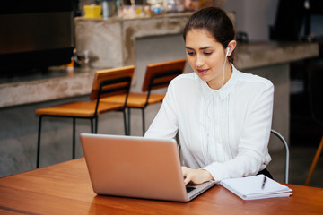 Young Caucasian woman working with laptop in coffee shop while listening with headphone