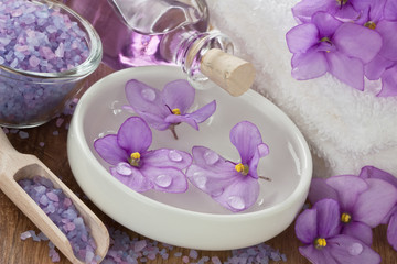 Fototapeta na wymiar SPA treatment set / Floating violet flower in water, aromatic essential oil, sea salt, soft towel and on wooden background