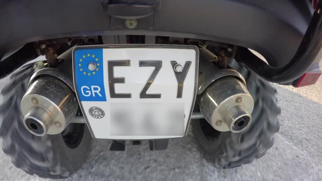 Action camera attached to rear of quad motor cycle showing rotating wheels, (blurred)license plate and exhaust pipes. Driving across a sunny road.