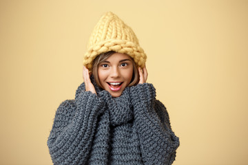 Young beautiful fair-haired girl in knited hat and sweater smiling looking at camera over yellow background.