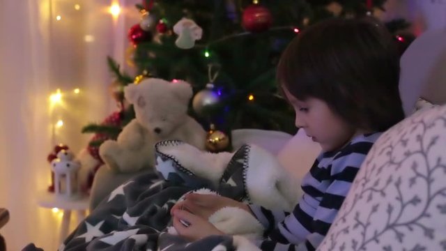 Cute sweet child, boy, playing on mobile phone under the blanket on first advent Sunday, christmas tree behind them.