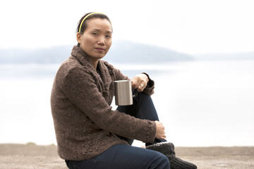 Asian woman enjoys coffee by lake.