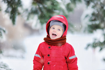 Portrait of funny little boy in red winter clothes having fun in snowfall