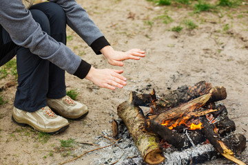  Woman  warms  hands over the fire, dressed in trekking clothes sitting beside a campfire. Active outdoor recreation,  holiday camping. 