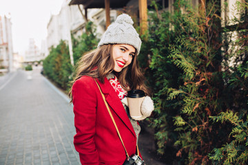 Pretty young girl with long hair in red coat and white gloves walking on street with coffee to go....