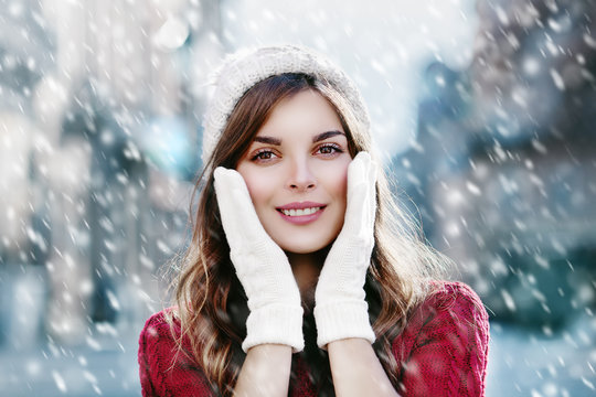 Young beautiful happy smiling girl walking on the street. Model looking at camera, touching her face, wearing stylish knitted hat and gloves. Christmas, New Year, winter holidays concept. Close up
