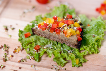 Slice of dark (brown) rye bread with liver pate, radish sprouts and paprika on a wooden cutting board. Checkered towel/pad. Delicious breakfast full of vitamins. Healthy lifestyle.