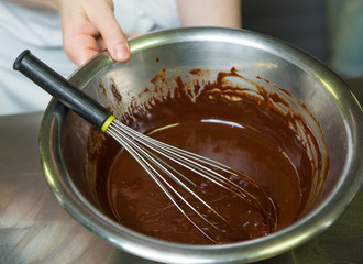 A metal bowl and whisk with rich thick dark melted chocolate sauce, in a metal industrial kitchen.
