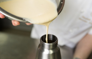 Thick fresh sweet dessert cream, being poured into a metal container in an industrial kitchen.